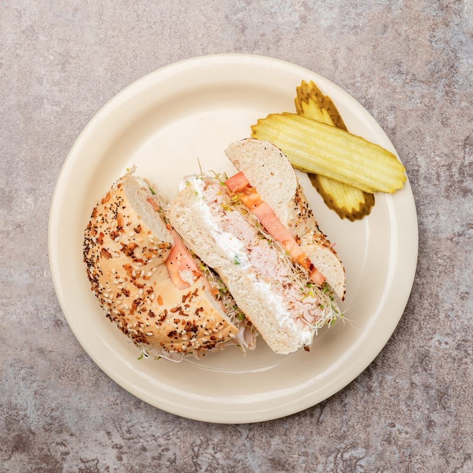 Everything bagel sandwich with cream cheese, sprouts, and tomato, served with pickle slices.