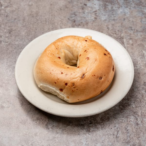 A plain bagel with a few dark specks sits on a white plate against a gray background.