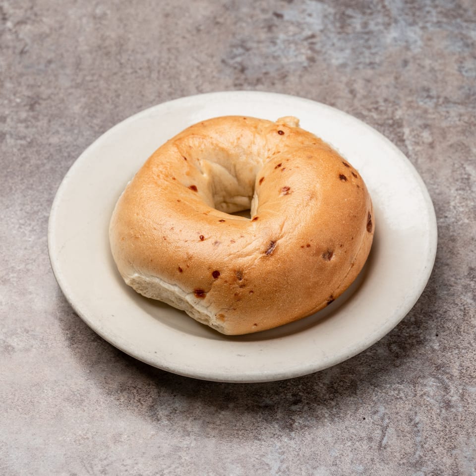 A plain bagel with a few dark specks sits on a white plate against a gray background.